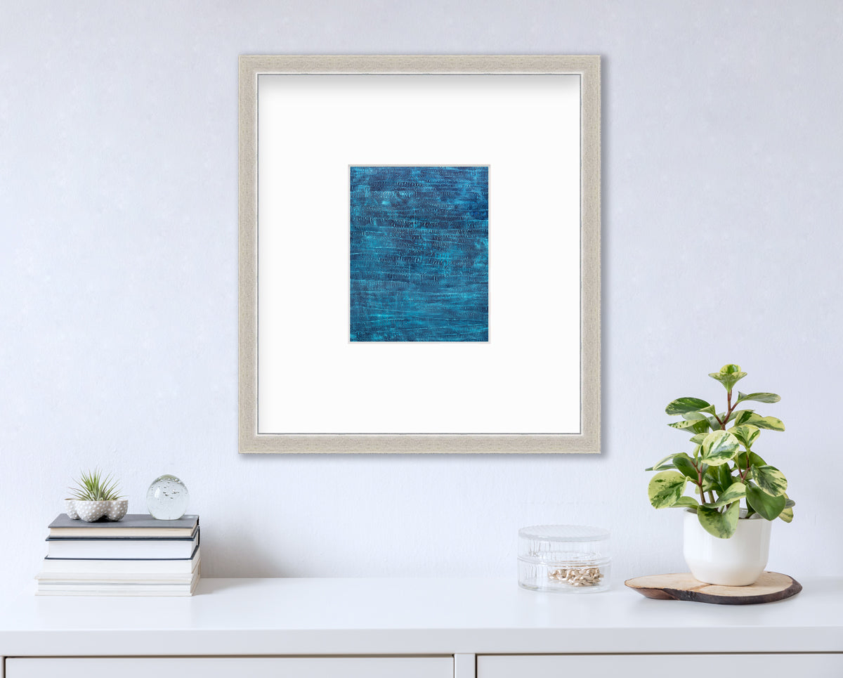 A dark teal abstract print in a silver frame with a mat hangs on a white wall above a white surface with a stack of books, a round clear container with gold accessories and a potted plant.