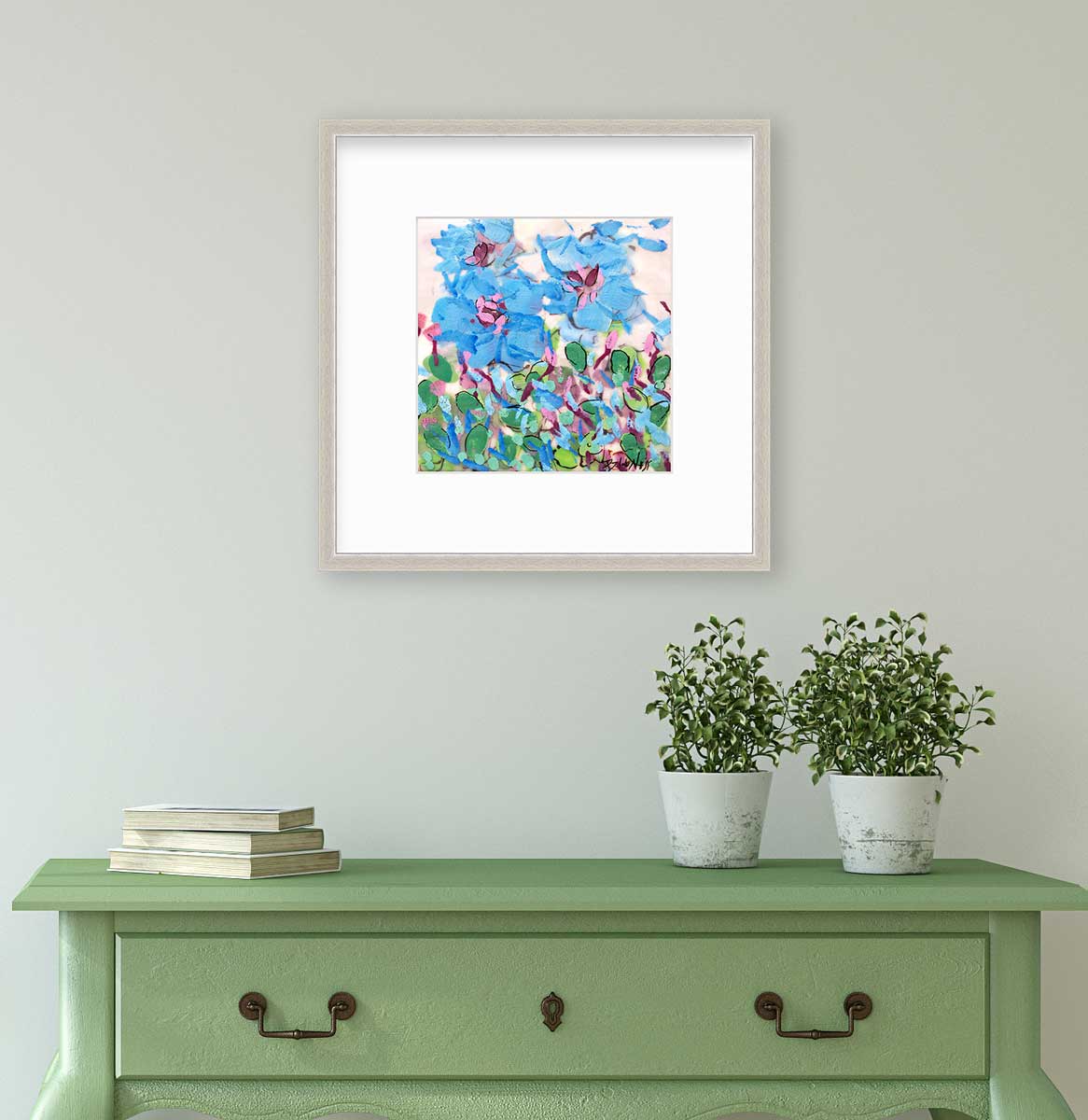 A blue, pink, green and white abstract floral print in a silver frame with a mat hangs on a white wall above a green console table with a stack of books and two potted plants.