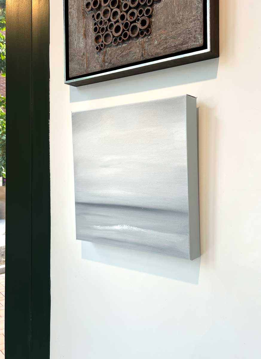 A right side-angle view of a tonal grey and white abstract painting by Tony Iadicicco hangs on a white wall.