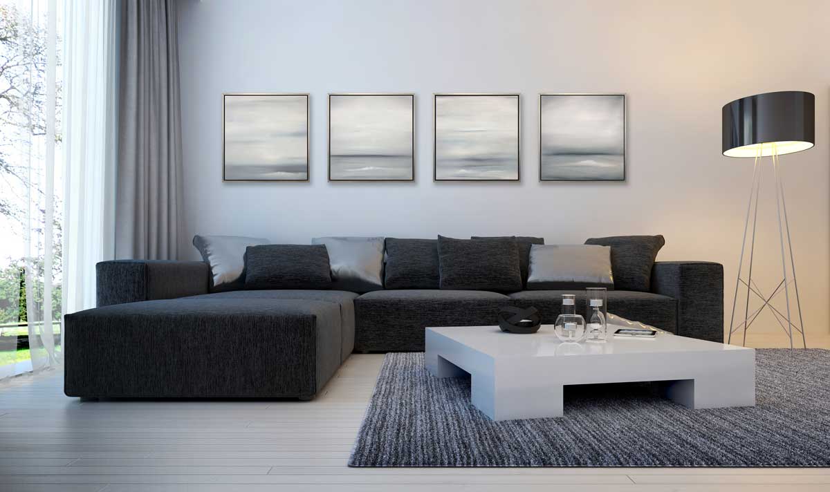 Four tonal grey abstract prints in silver floater frames hang side by side on a white wall above a black sectional sofa with pillows, an oversized grey square coffee table and a floor lamp with a black shade.