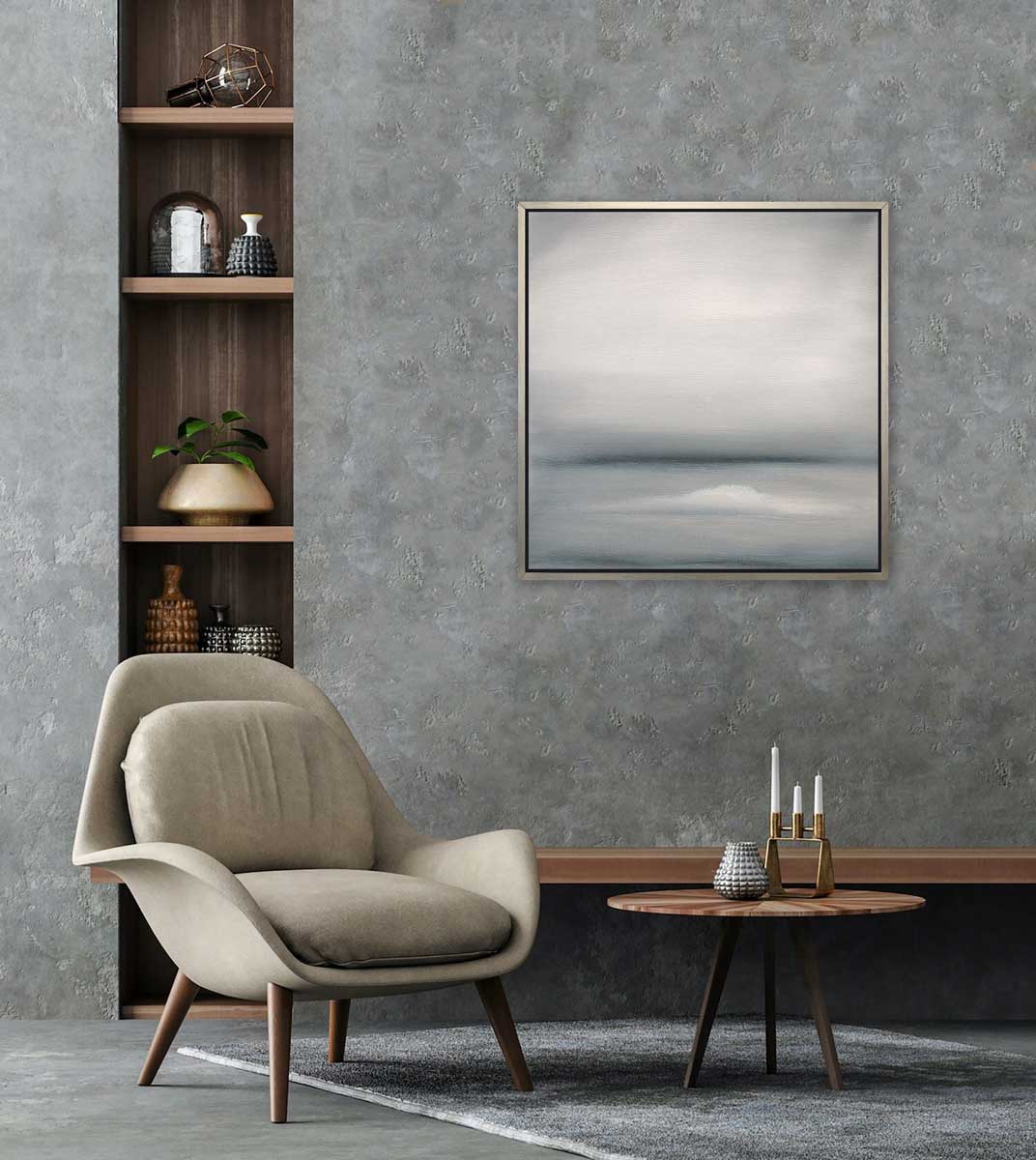 A tonal grey abstract print in a silver floater frame hangs on a grey textured wall above an off-white modern chair, a round wood side table and a built-in vertical bookshelf.