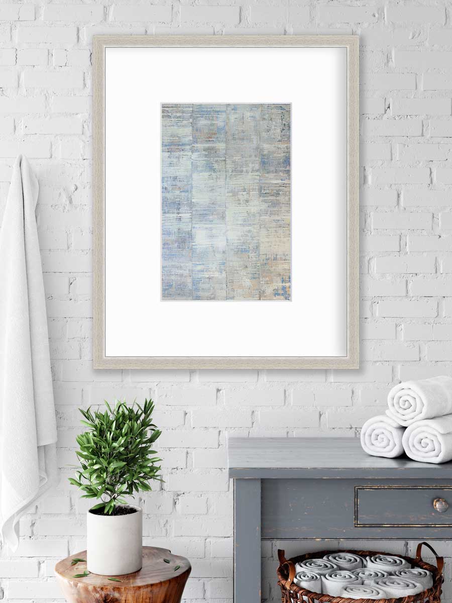 A white and blue abstract print in a silver frame with a mat hangs on a white wall above a grey side table with rolled towels, a basket underneath with more rolled towels and a side table with a potted plant.