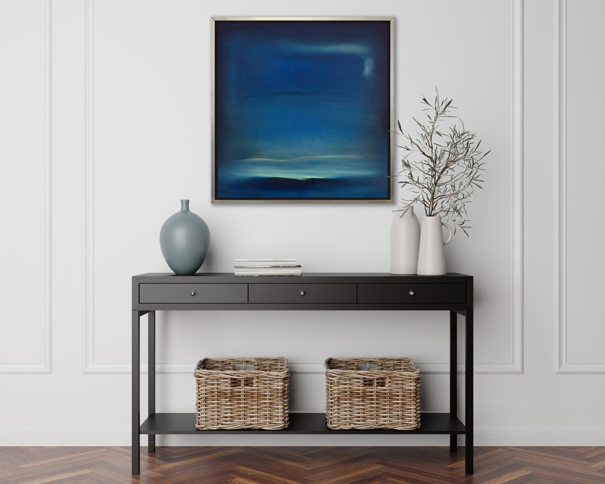 A dark blue and teal with accents of light yellow abstract print in a silver floater frame hangs on a white wall above a black console table with two greenery filled white vases, a muted teal vase, a stack of books and two wicker baskets.