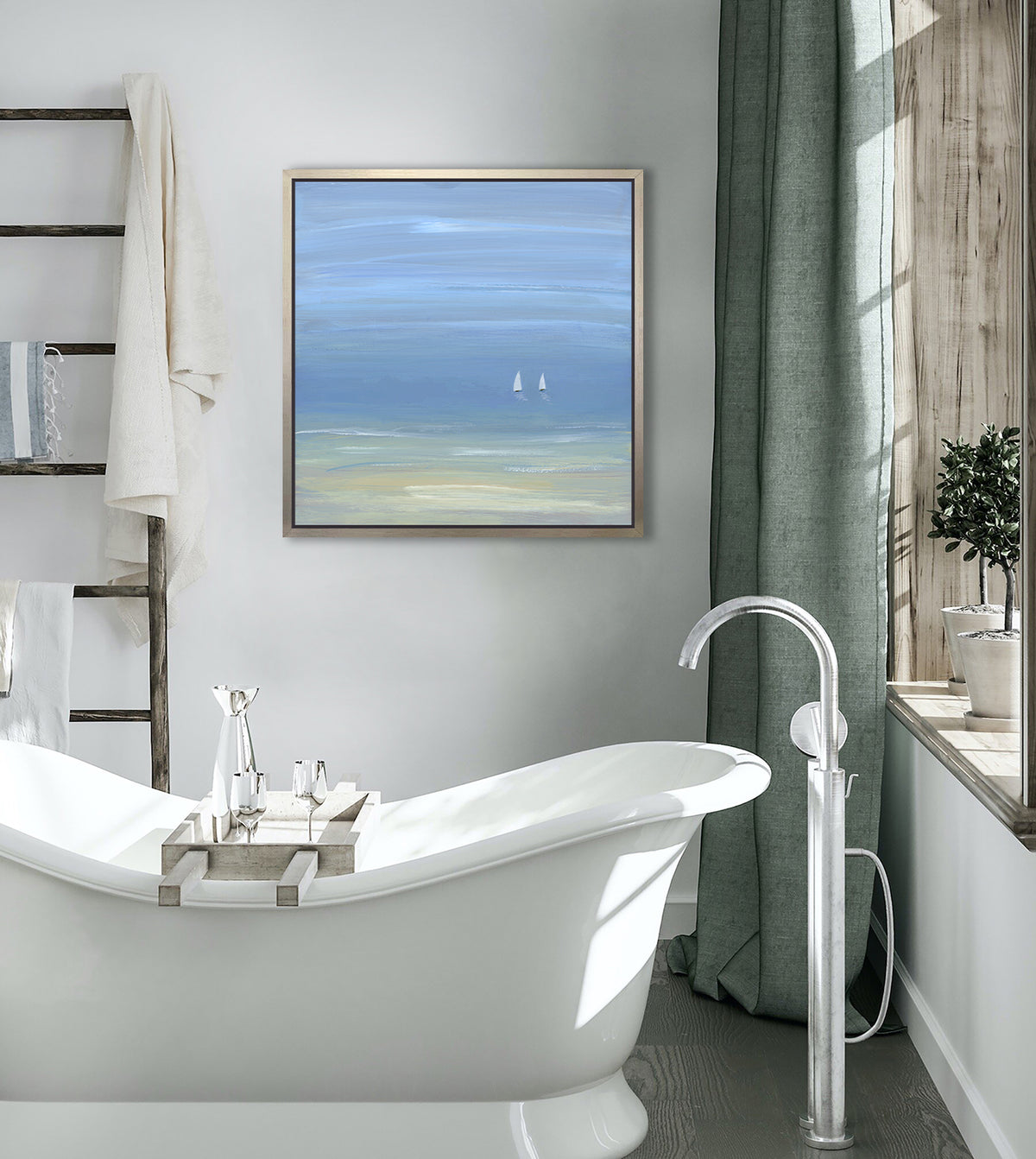 A blue, beige and white abstract seascape print with two small sailboats in a silver floater frame hangs on a white wall above a modern white soaking bathtub, a ladder holding towels and a window sill with potted topiaries.