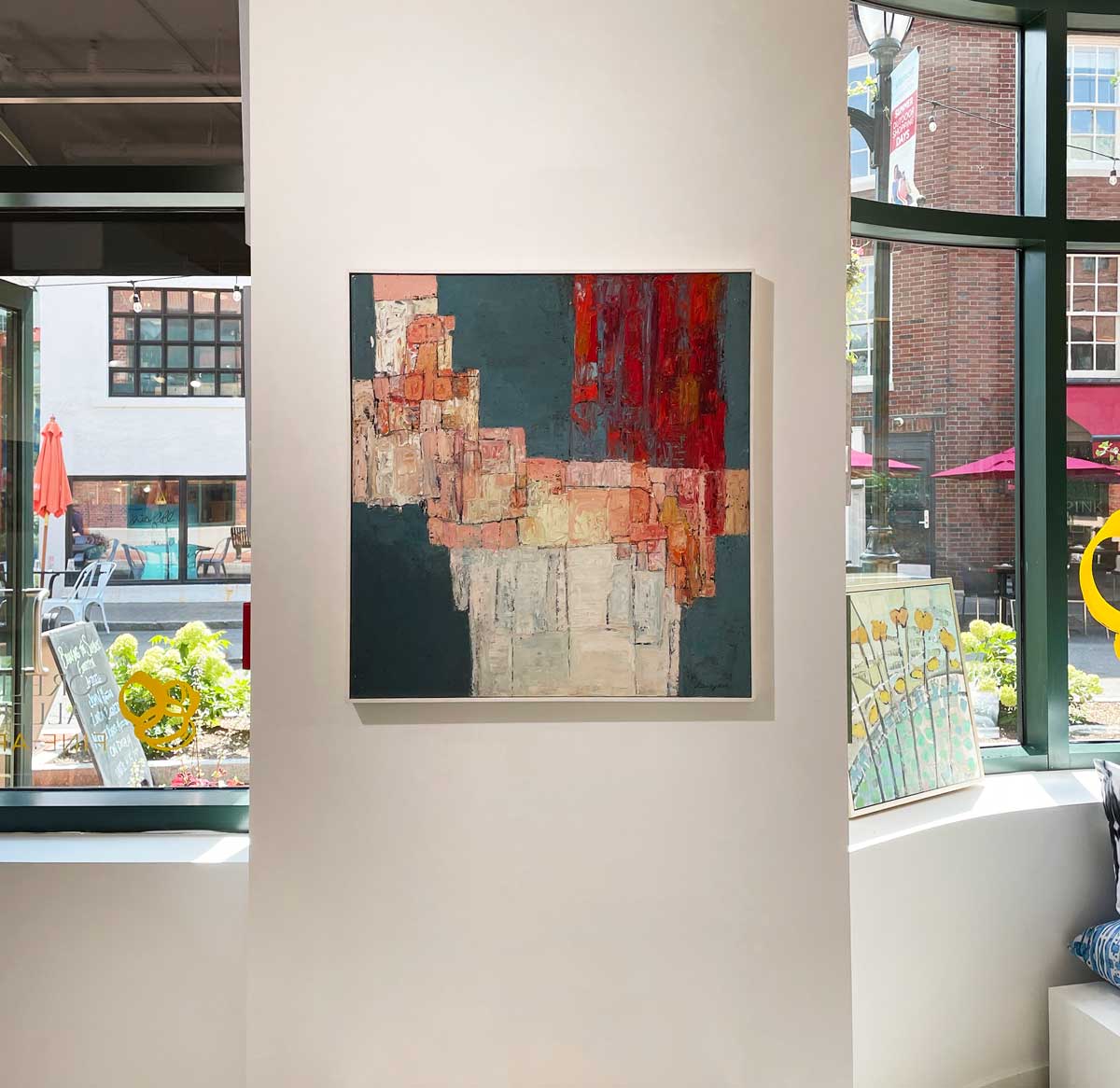 A red, dark teal, pink and white abstract painting in a white frame hangs on a white wall.