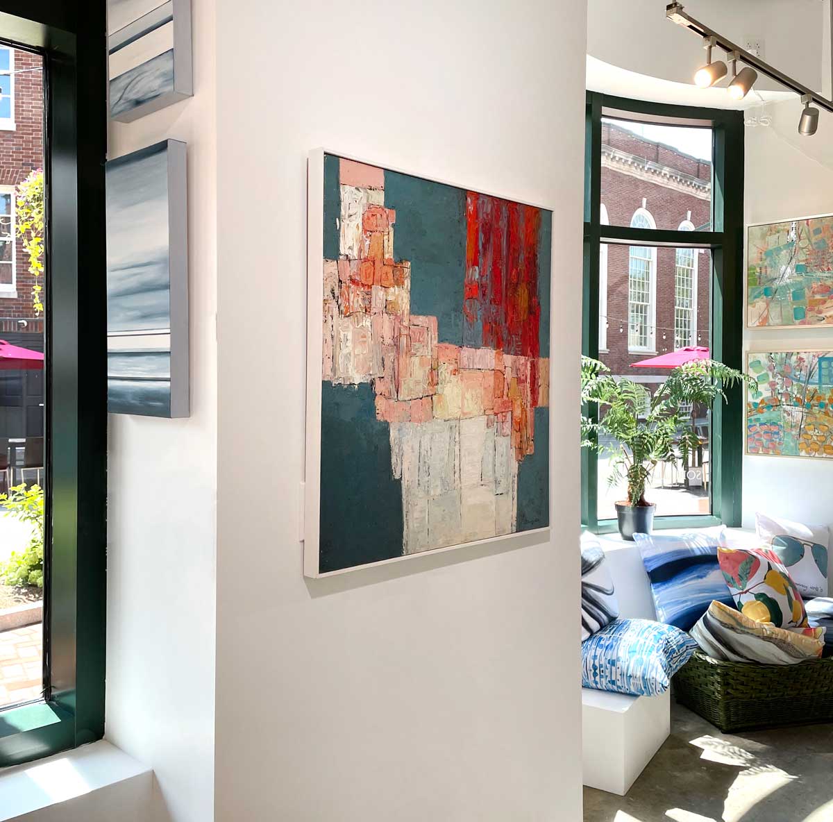 A left side-angle view of a dark teal, pink, white and red abstract painting in a white frame hangs on a white wall.