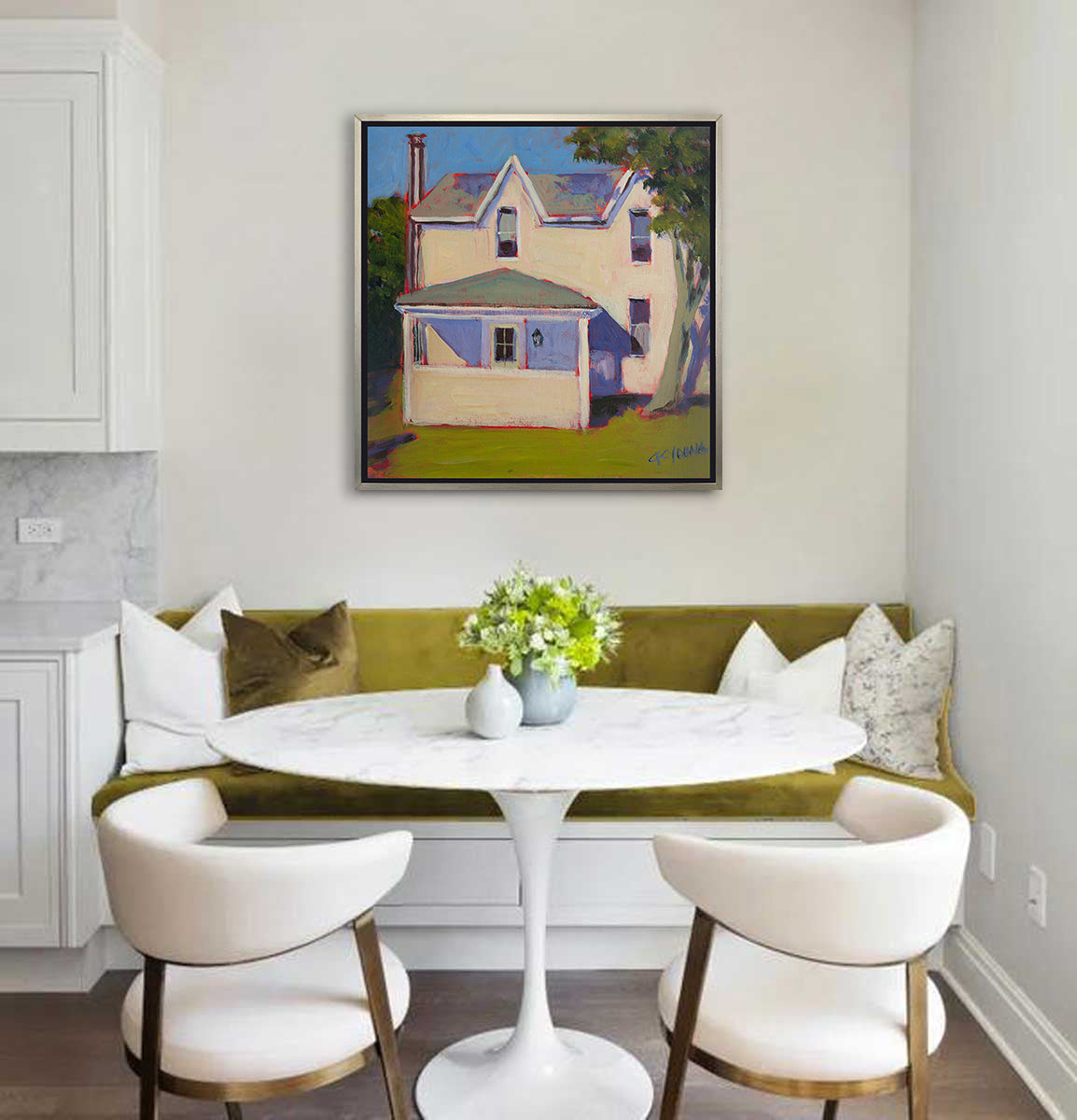 A print of a white house that sits next to a giant tree, green grass in front in a silver floater frame hangs on a white wall above a chartreuse booth seat with pillows, a white round marble table and two white chairs.