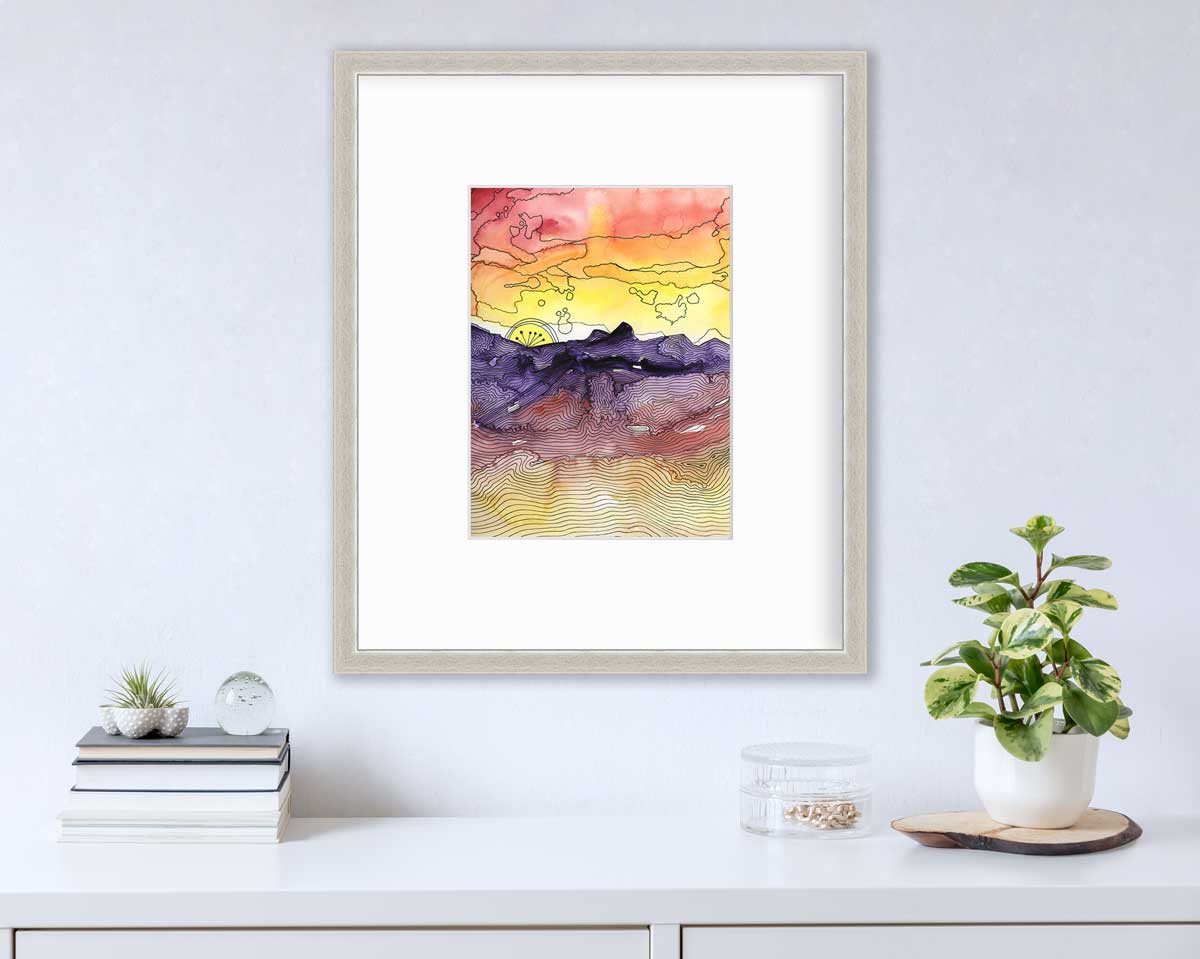 A red, orange, yellow, purple and black abstract landscape print in a silver frame with a mat hangs on a white wall above a white surface with a stack of books, a clear round container with gold accessories and a potted plant.