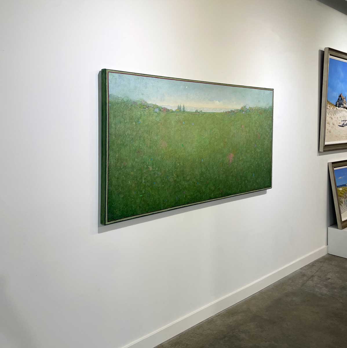 A left side-angle view of a green and blue abstract landscape painting by Elwood Howell hangs on a white wall.