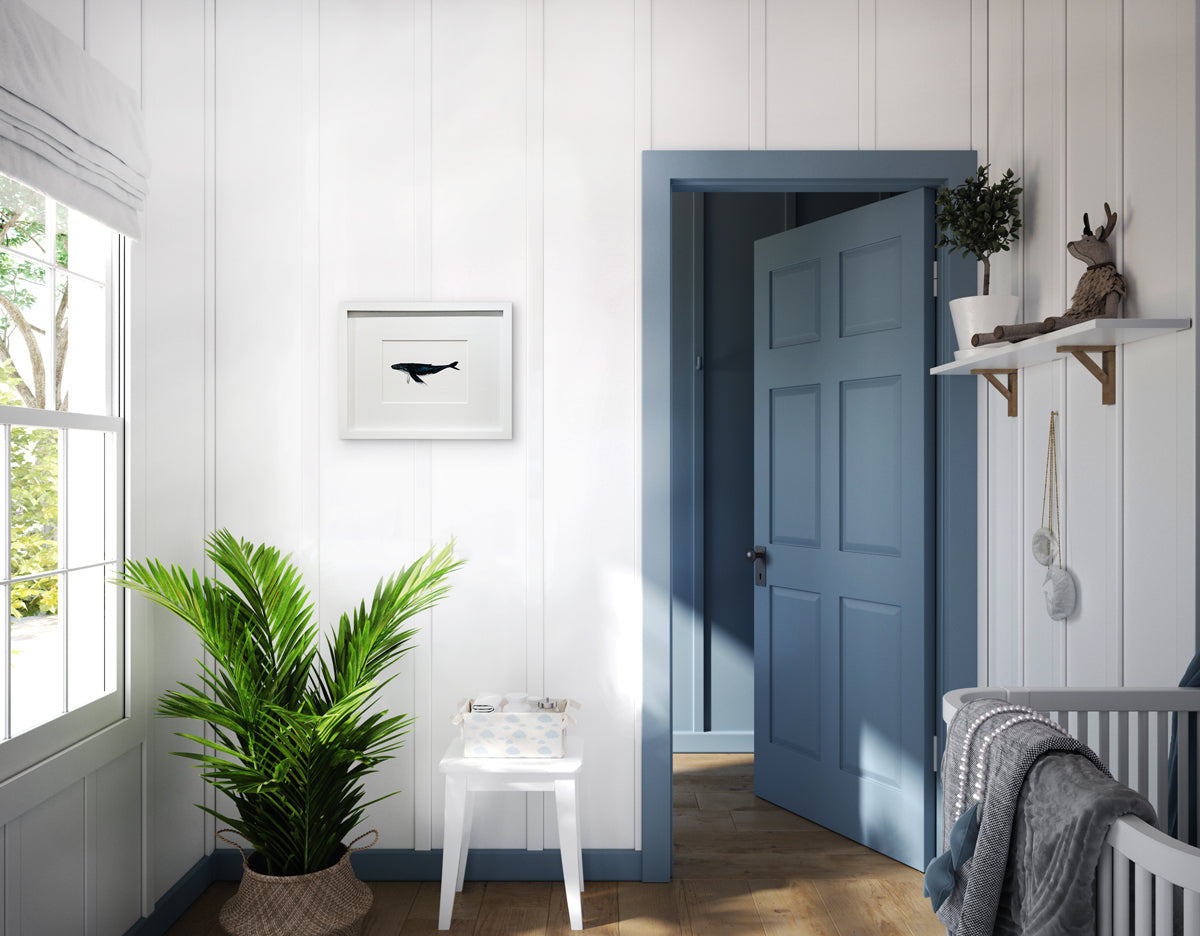 A blue, white and black illustration drawing of a whale in a white frame with a mat hangs on a white wall above a potted floor plant and a white stool with a basket.