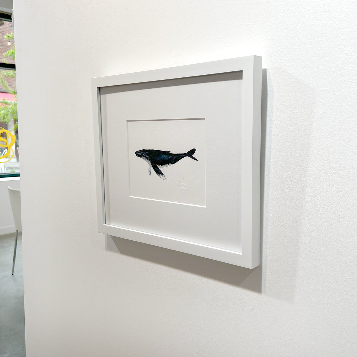 A right side-angle view of a blue, white and black illustration drawing of a whale in a white frame with a mat hangs on a white wall.