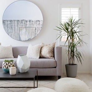 Circular mirror, mounted on wall, with grey mixed media painting on lower half. Displayed in living room above couch. Coffee table in front of couch and potted plant placed to the right of the couch with floor pillows in the foreground. 