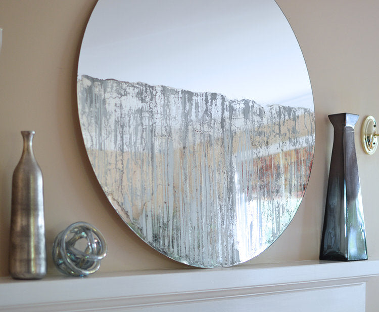 Circular mirror with grey mixed media painting on lower half. Displayed on the mantle, leaning up against the wall. Vases sit to the left and right of the mirror.
