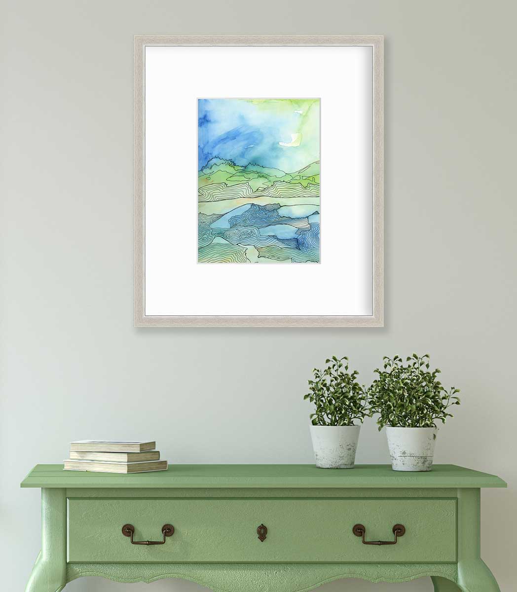 A green and blue abstract landscape illustration print in a silver frame with a mat hangs on a white wall above a green console table with a stack of books and two potted plants.