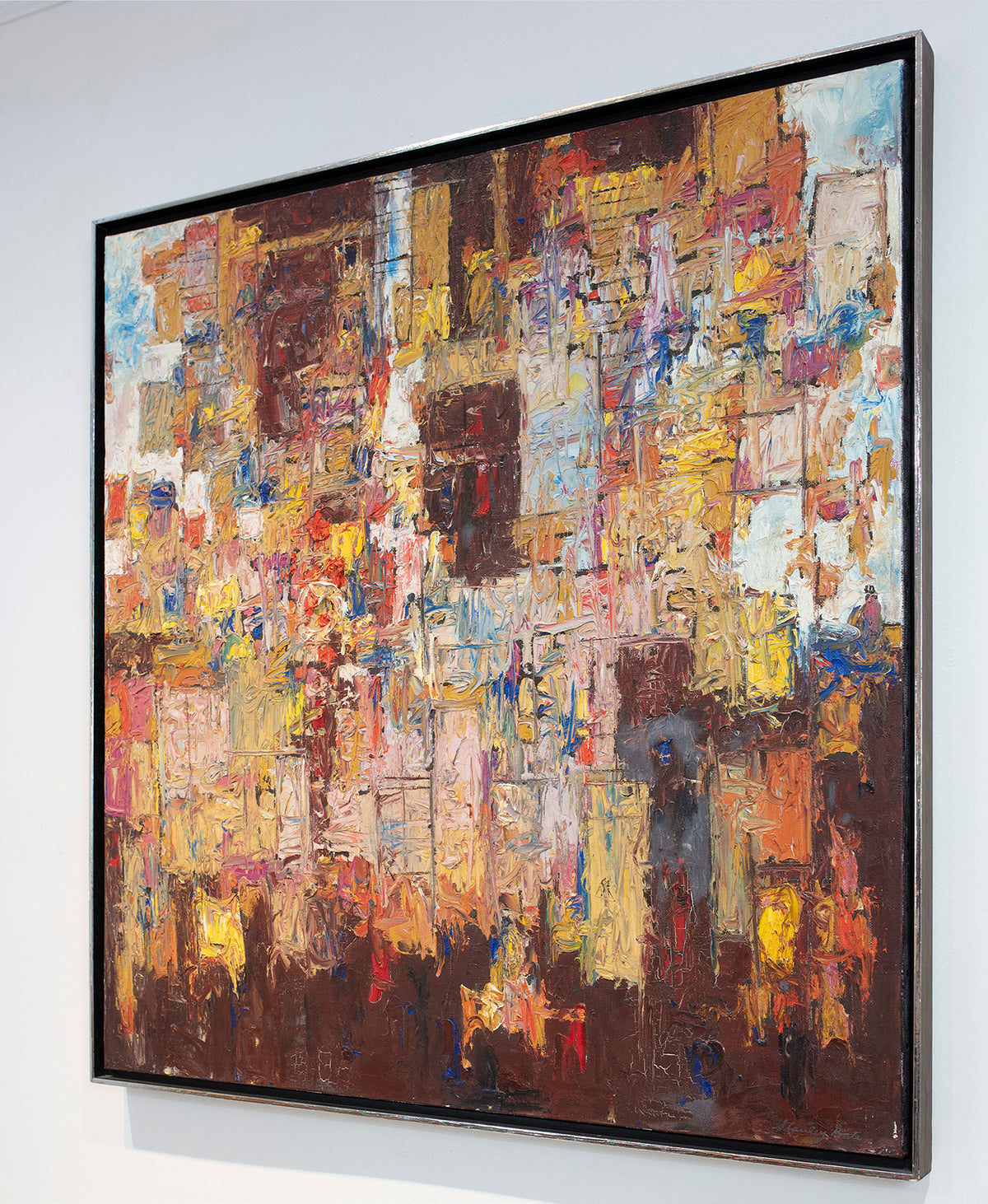A right side-angle view of a multicolored abstract modern painting in a silver frame hangs on a white wall.
