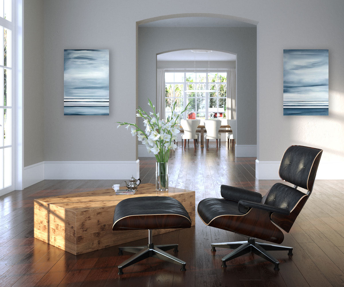 Two abstract paintings hang on a grey wall on either side of an archway that leads into a dining room. In the foreground sits a dark lounge chair and wooden coffee table.