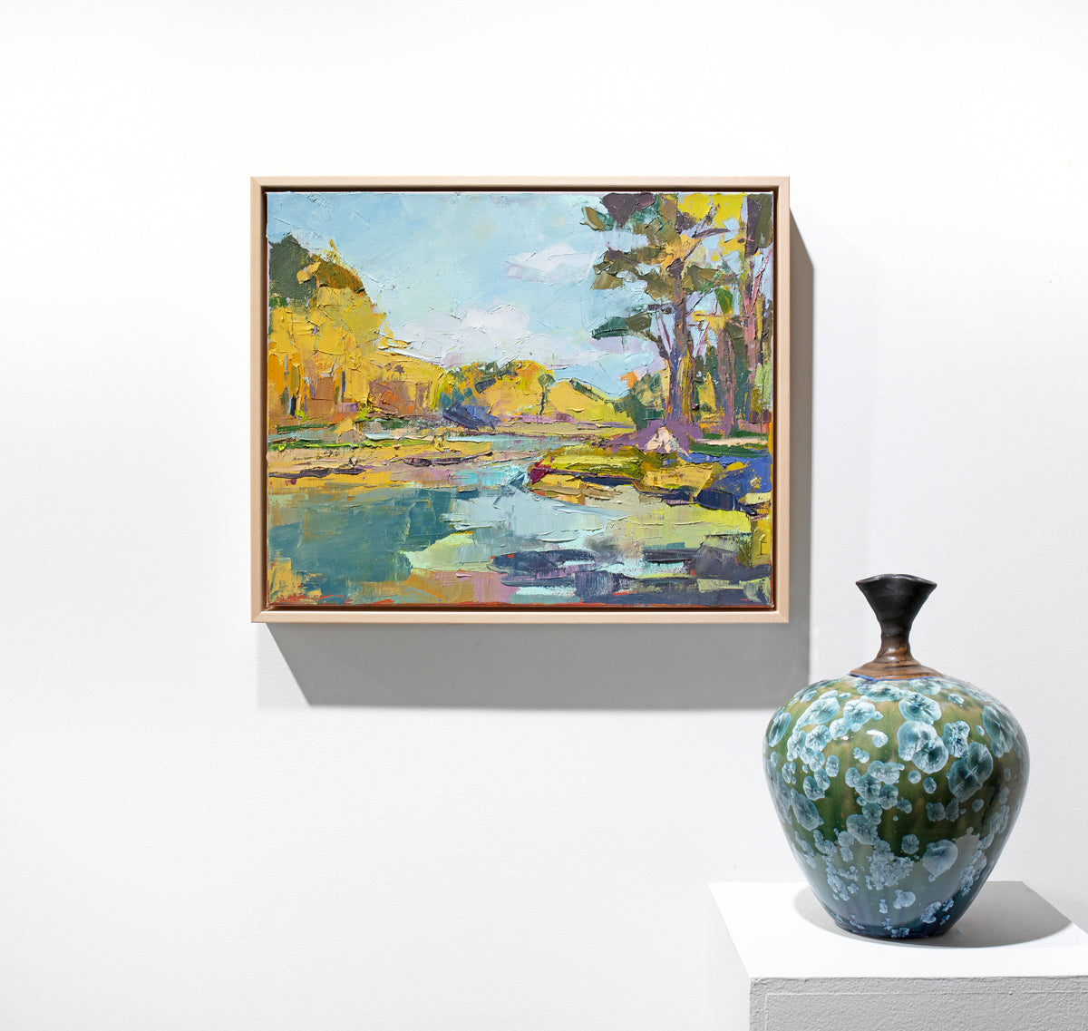 A blue and yellow abstract landscape painting hangs on a white wall with a teal and apple green ceramic pot to the right.