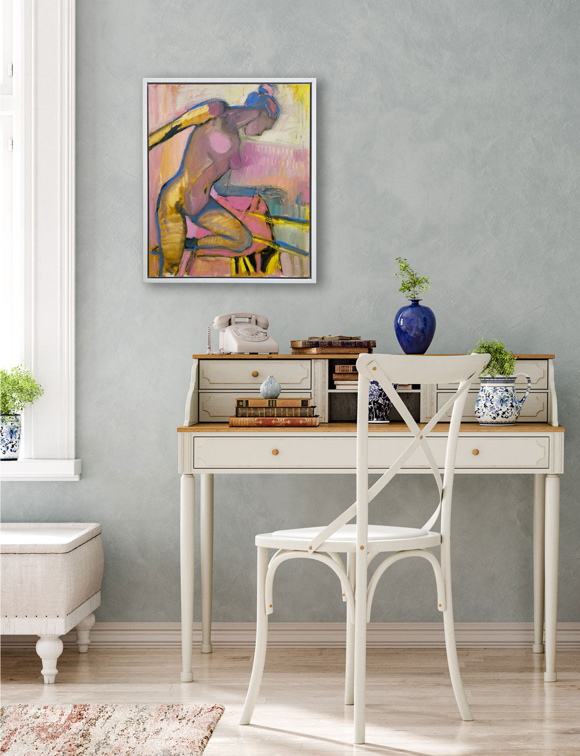 A framed yellow and pink abstract figurative painting hangs on a grey wall above a traditional desk and white chair next to a window.