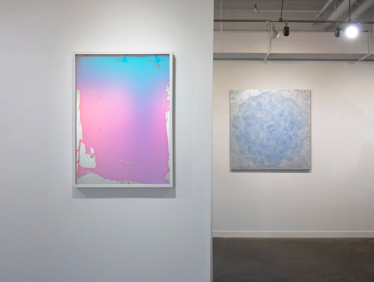 A pink and blue fine art mirror hangs on a wall, with an abstract painting hanging on a wall in the background beneath bright interior lights.