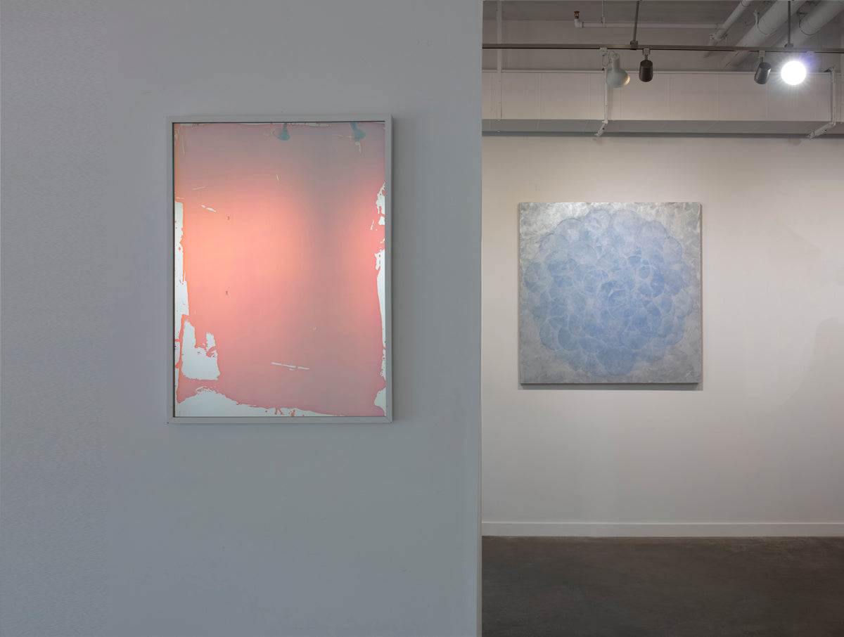A pink and blue fine art mirror hangs on a wall, with an abstract painting hanging on a wall in the background beneath bright interior lights.
