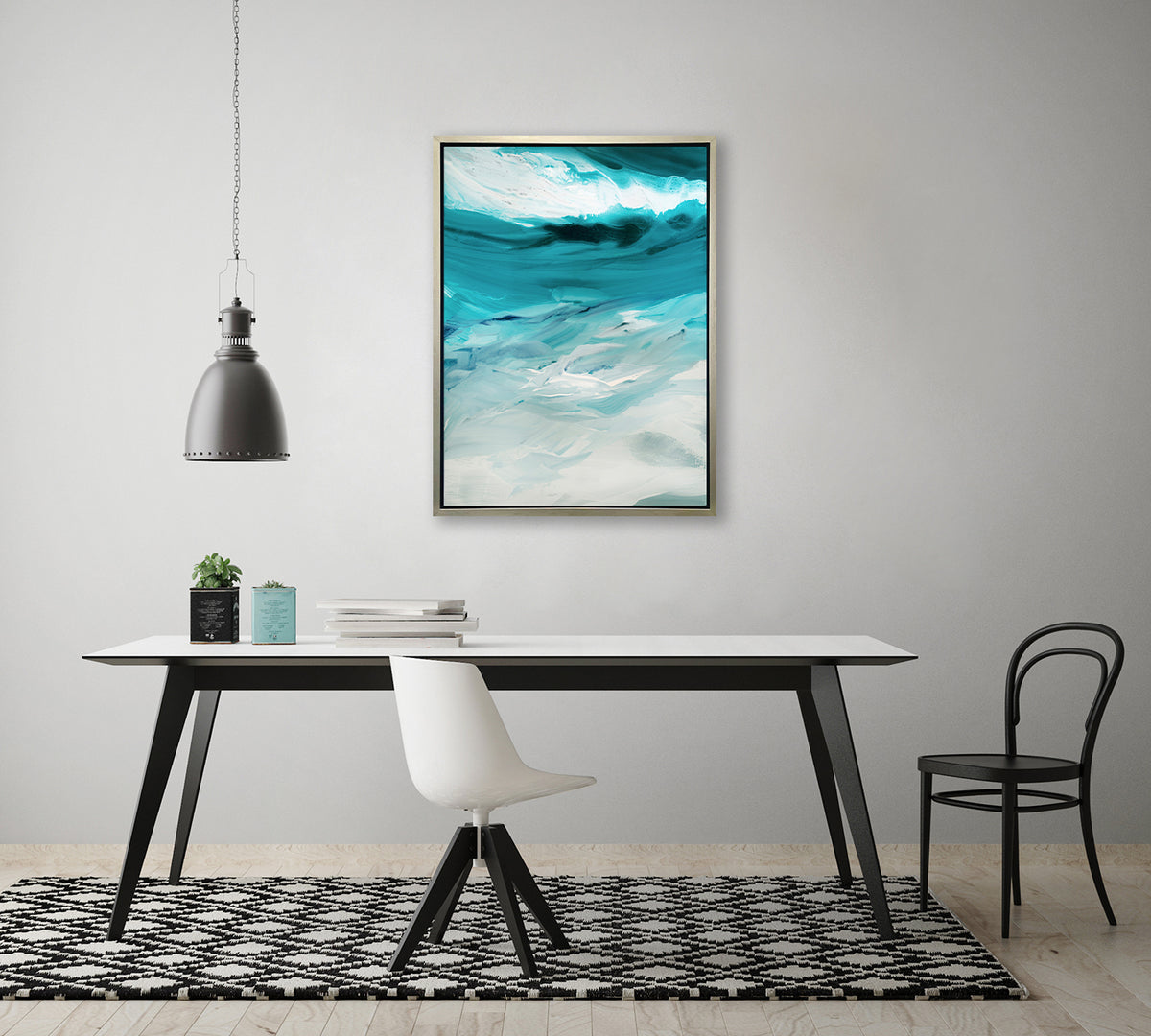 A teal, white and grey abstract print in a silver floater frame hangs on a white wall above a white table with a stack of books, a white chair and a black chair.