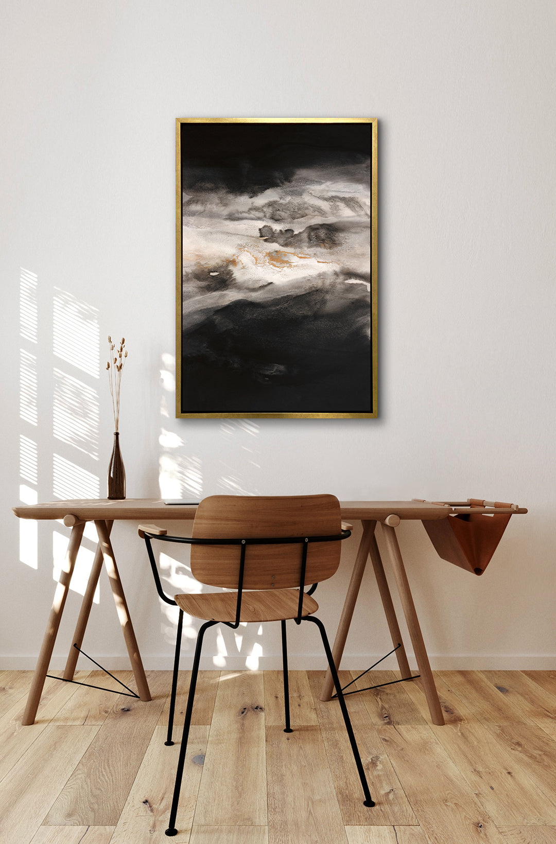 A black, white, beige and gold abstract print in a gold floater frame hangs on a white wall above a wood desk with a brown glass bottle and a wood and metal chair.