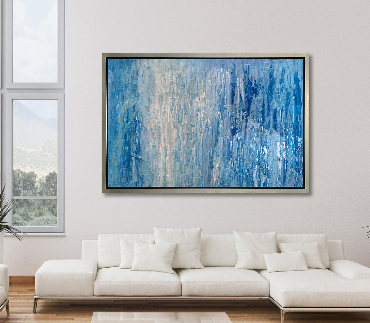 A blue, white and grey abstract print in a silver floater frame hangs on a white wall above a white sectional sofa with white pillows.