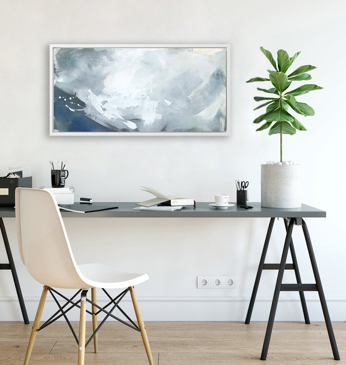 A blue, grey and white abstract print in a white floater frame hangs on a white wall above a grey desk with a open book, a black mug with pencils and a potted plant.