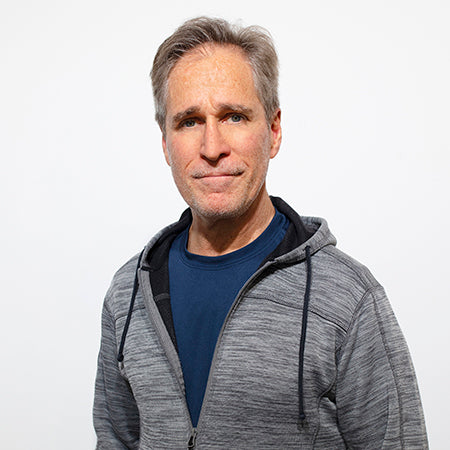 Headshot of artist, Joe Sorge, wearing blue shirt and grey hoodie, and standing in front of a white wall. 