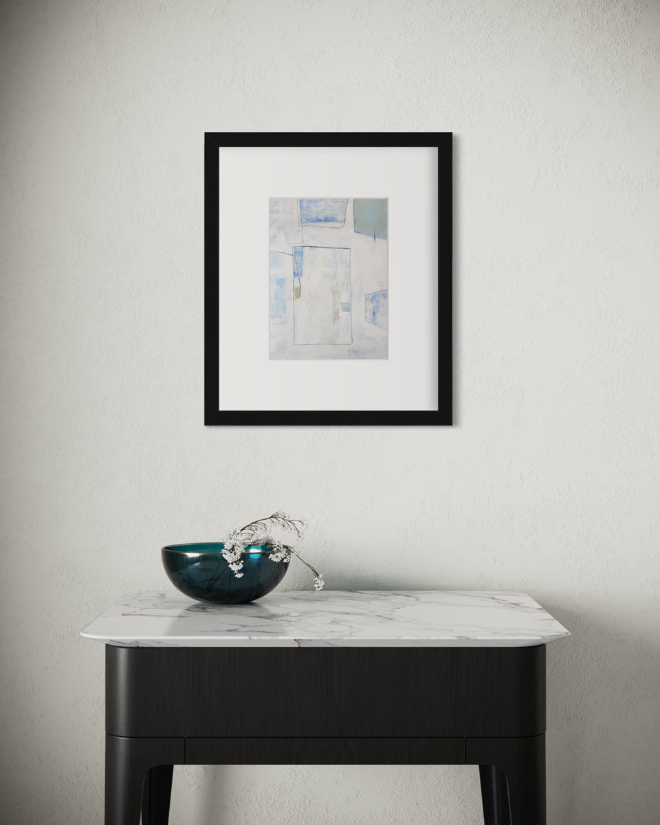 A white, blue and green abstract print in a black frame with a mat hangs on a white wall above a black side table with a white marble top and a teal bowl with white flowers.
