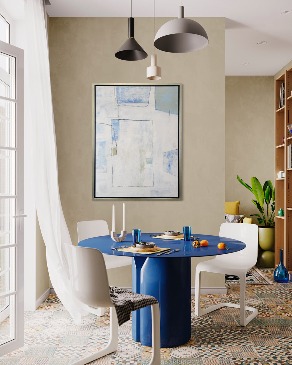 A white, blue and green abstract print in a silver floater frame hangs on a beige wall above a round blue pedestal table with place settings and three white chairs.