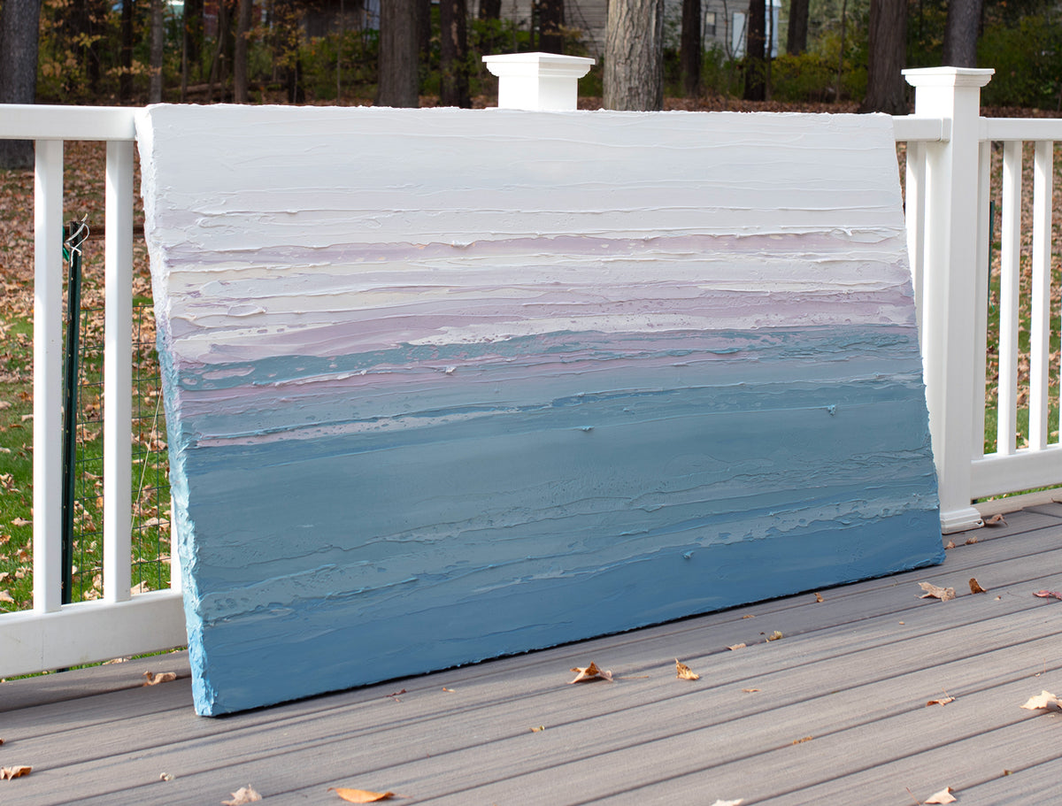 A white, lavender, teal and blue abstract heavily textured painting like sculpture by Teodora Guererra leaning outside in natural light on a deck.