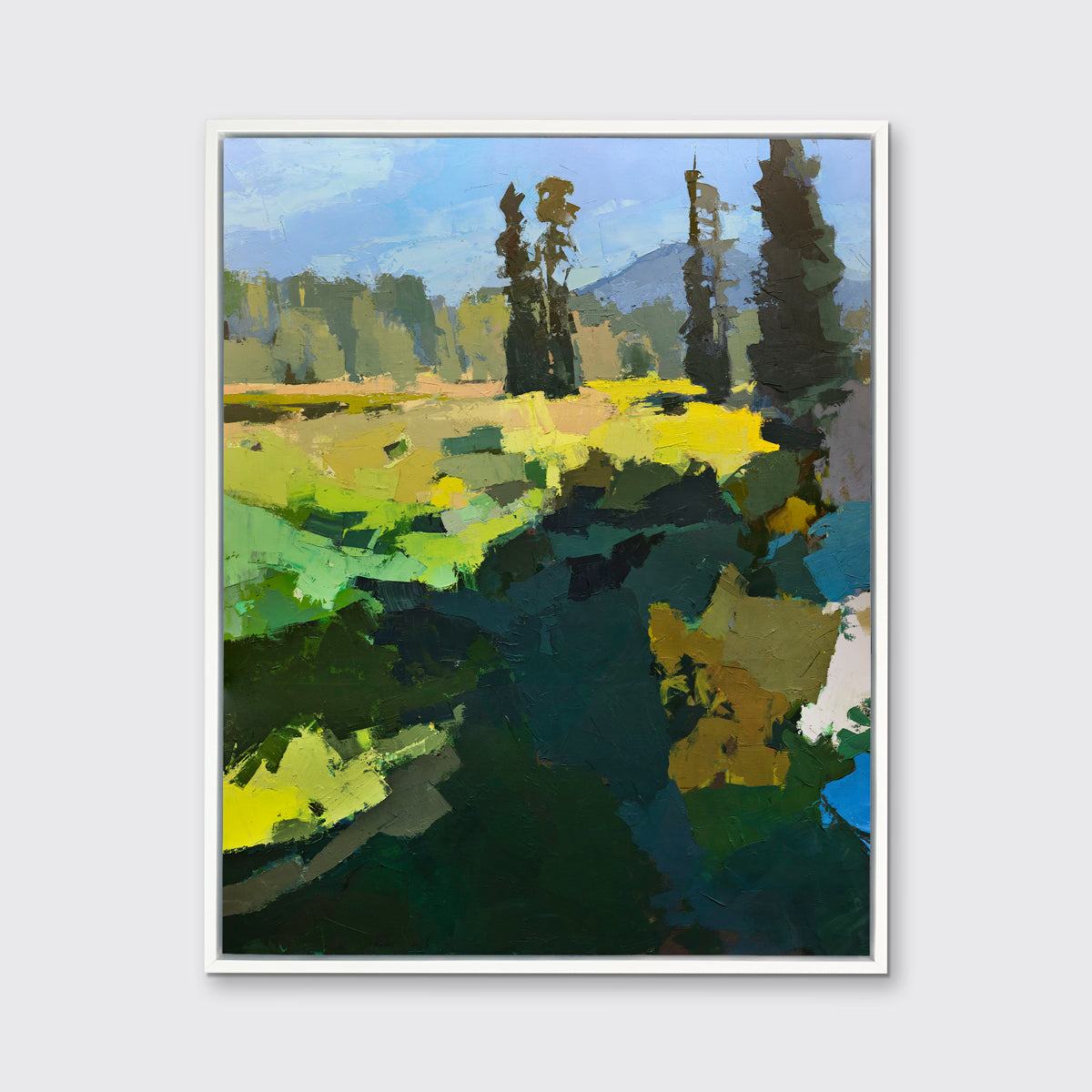 A blue, green, and yellow abstracted landscape art print framed in a white frame hangs on a white wall.