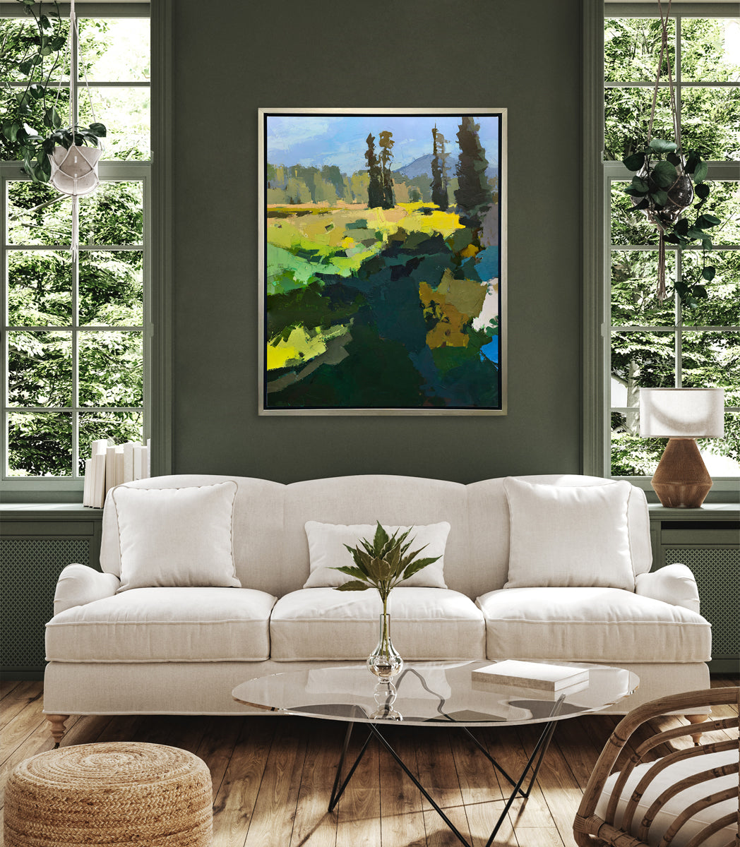 A blue, green, and yellow abstracted landscape art print framed in a silver frame hangs on a green wall above a white sofa and between two windows. 
