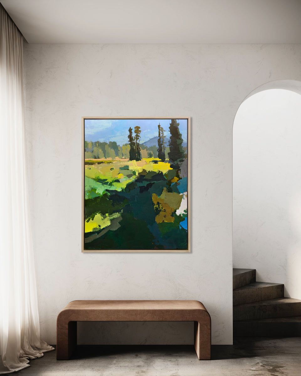 A blue, green, and yellow abstracted landscape art print framed in a white oak frame hangs on a white wall above a modern wooden bench and next to an archway with a staircase. 