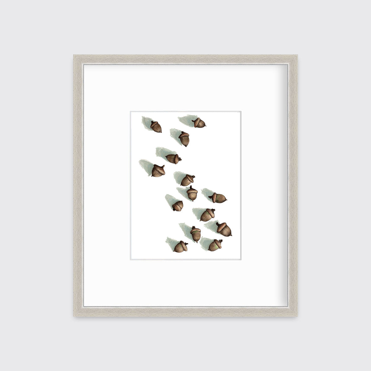 A print of several brown acorns with long shadows on a white background in a silver frame with a mat hangs on a white wall.