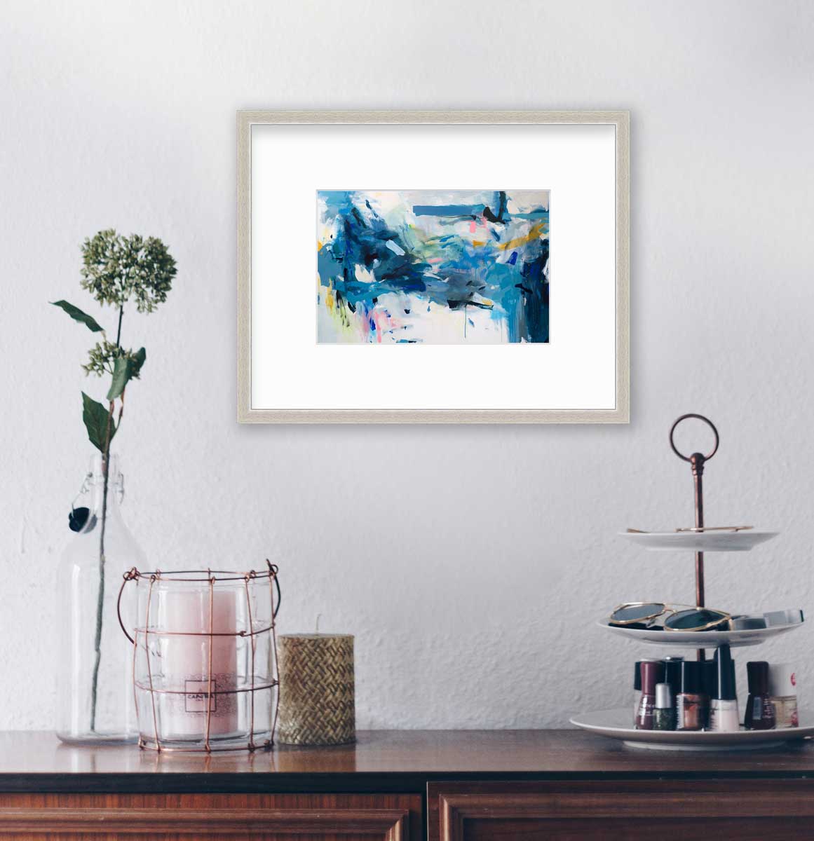 A blue and white abstract print by Kelly Rossetti in a silver frame with a mat hangs on a white wall above a wood surface with a three-tiered caddy with nail polish, two candleholders and a clear bottle with a floral stem.