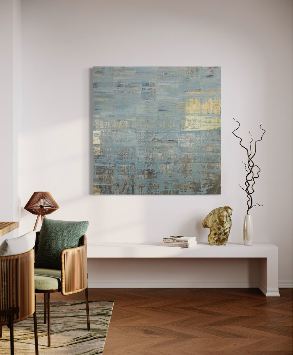 A blue and gold abstract painting hangs on a white wall over a white bench with books, gold sculpture and vase with decorative twigs.