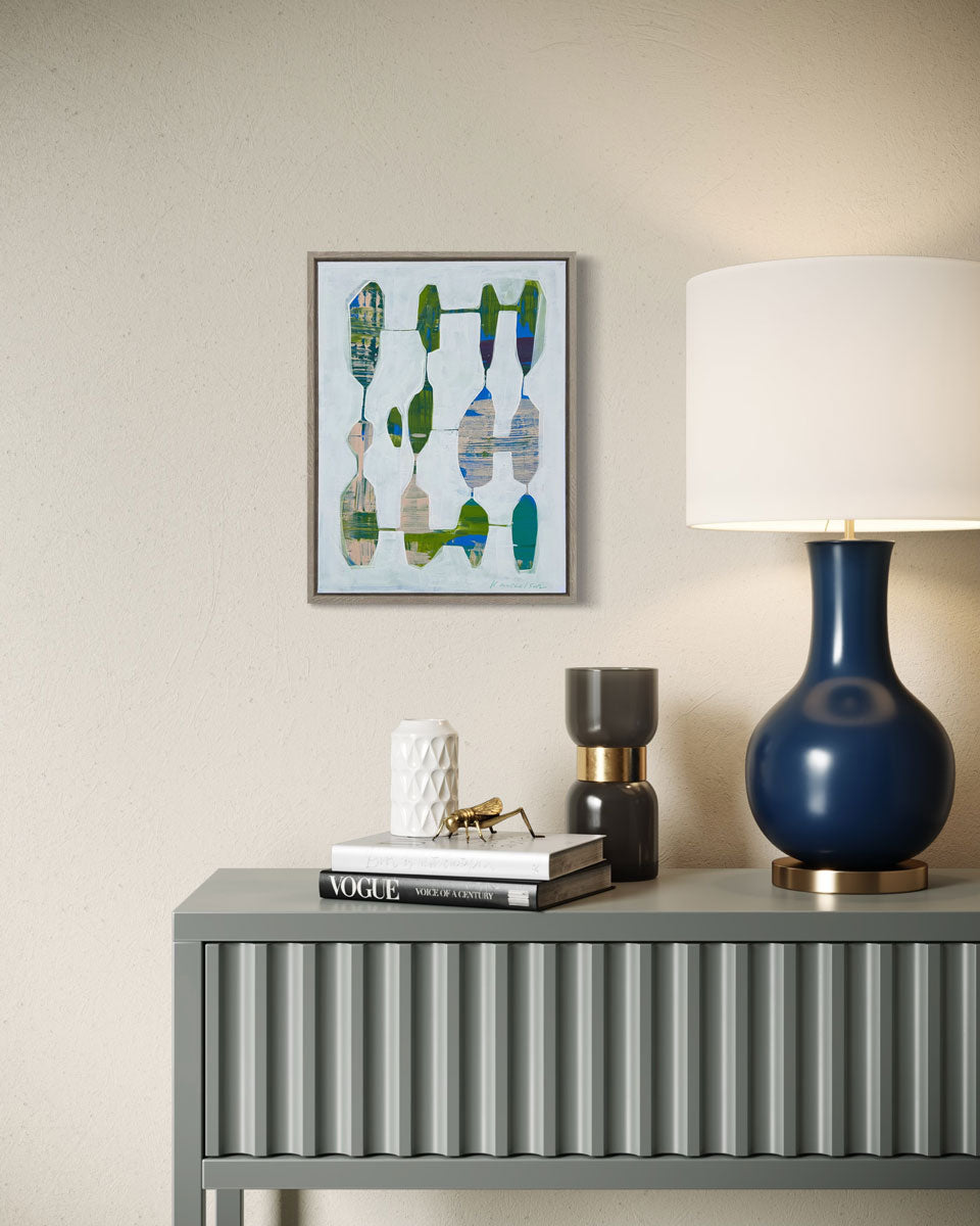 A small framed abstract painting hangs on a light wall above a modern console table with modern table decor and a blue lamp. 