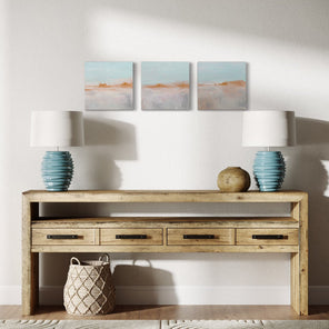 Three abstract paintings of a pink and grey landscape with a golden horizon and light blue sky hanging on a white wall over a natural wood farmhouse credenza with 2 ceramic teal lamps and a clay pot. 