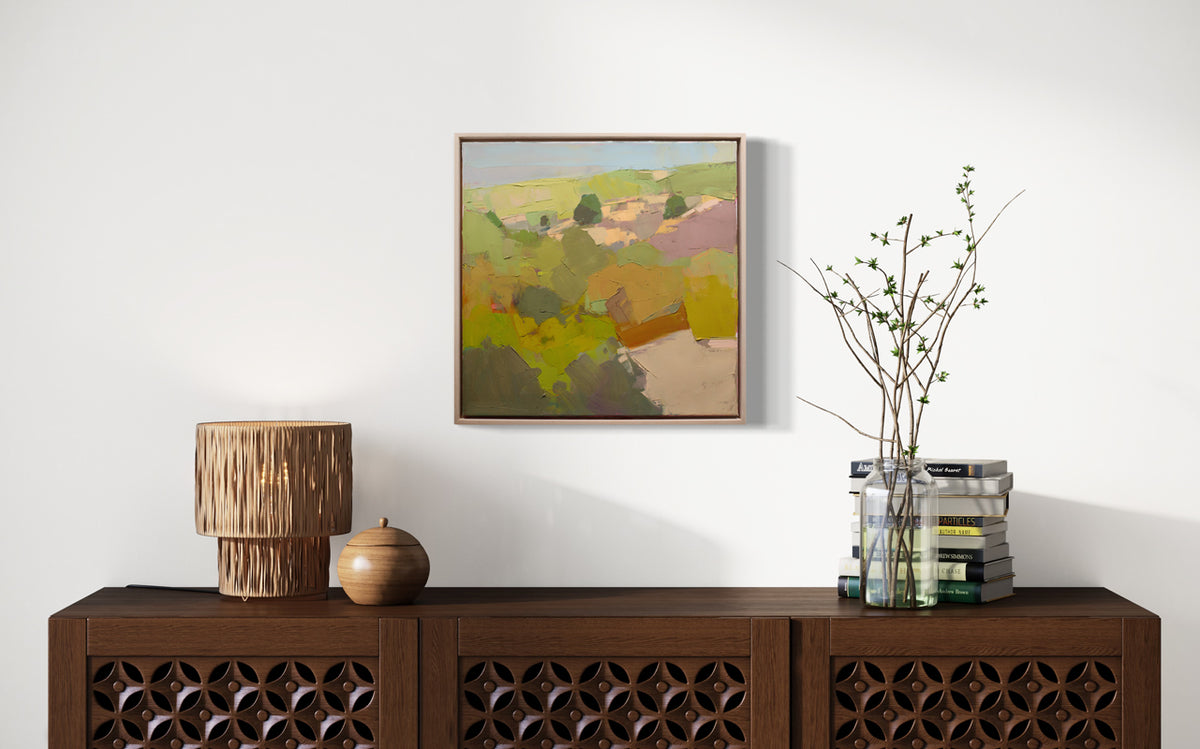 A green abstracted landscape painting framed in a wood floating frame hangs on a white wall above a wood console table with wood table decor and books. 