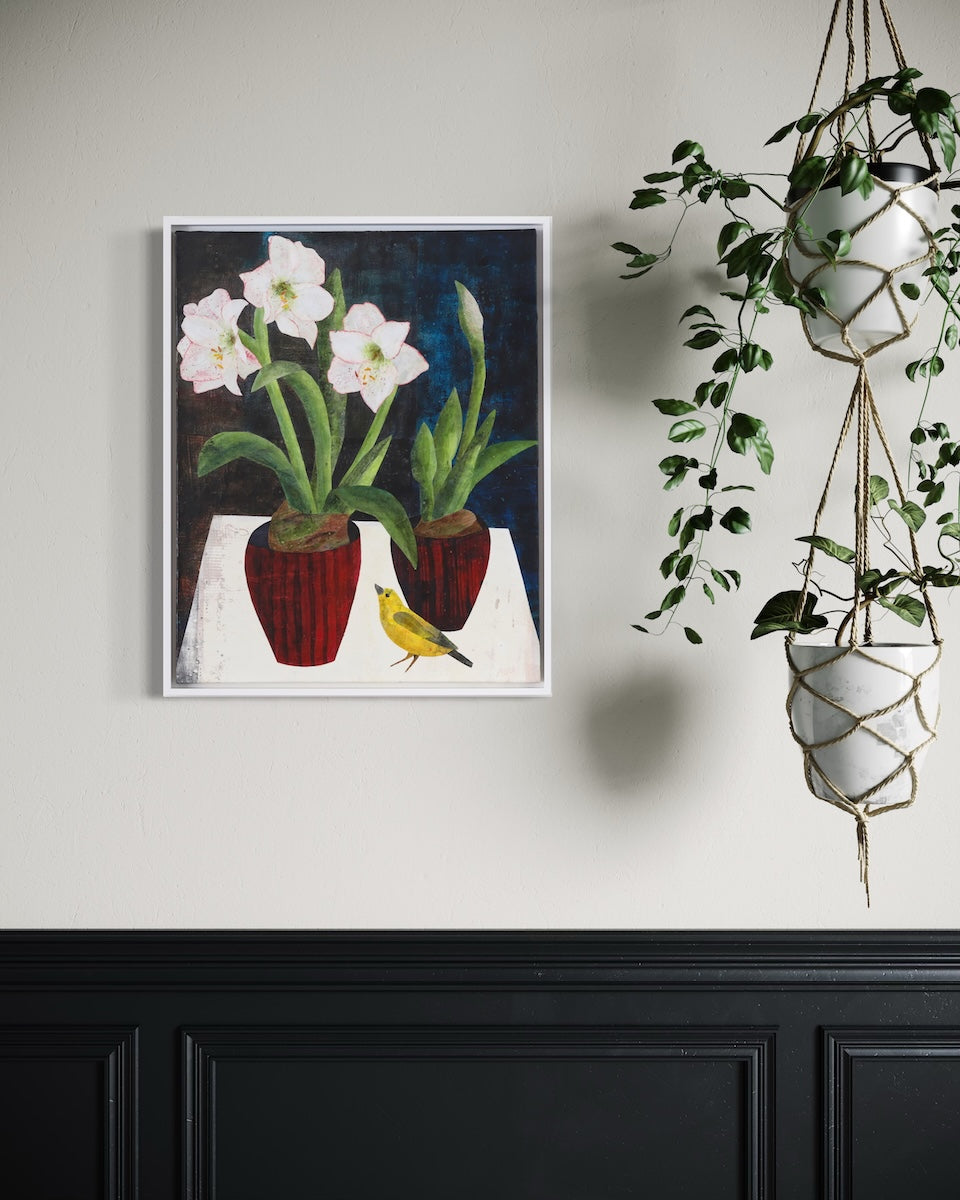 A framed floral collage hangs on a white wall next to a hanging potted plant. 