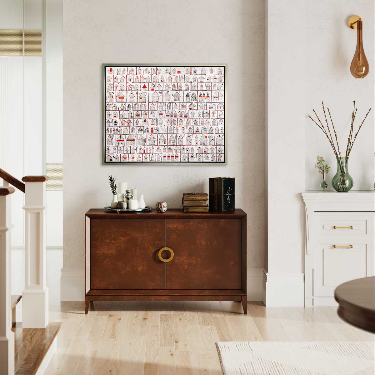 A red and white abstract house print in a silver floater frame hangs on a white textured wall above a dark wood console cabinet with decorative accessories and next to a staircase.