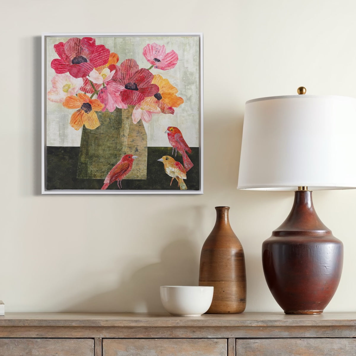 A framed square collage depicting pink and orange flowers in a vase with small birds, displayed on a beige wall above a console table with a lamp, vase, and bowl.