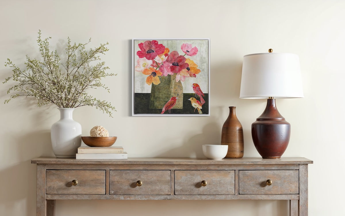 A framed floral and bird painting hangs on a beige wall above a rustic wooden console table styled with a white vase of branches, books, a bowl, a ceramic lamp, and other decorative objects.