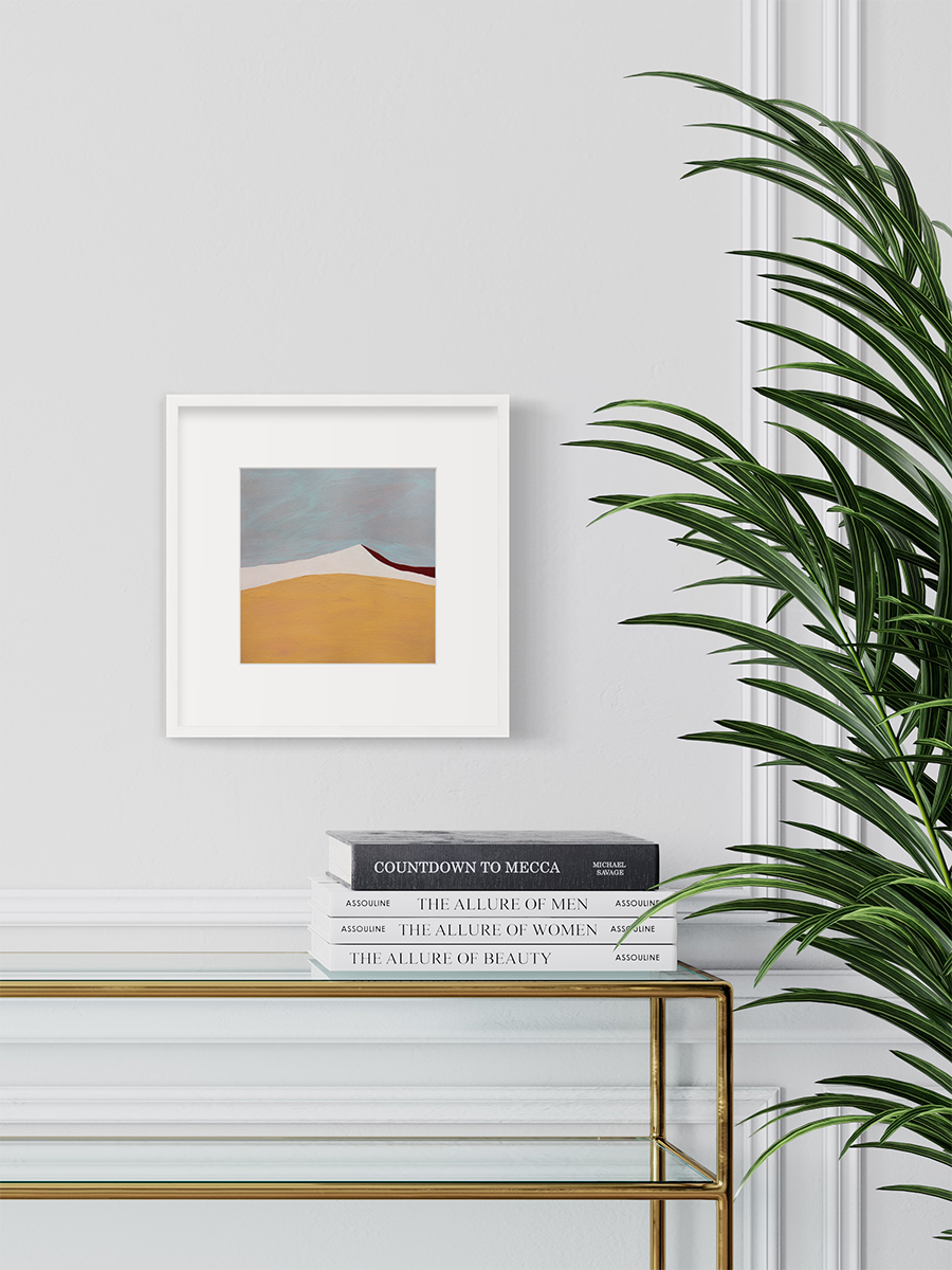 A dark yellow, white, dark red and slate blue abstract landscape print in a white frame with a mat hangs on a white wall above a gold console table with a stack of books and a large potted plant to the right.