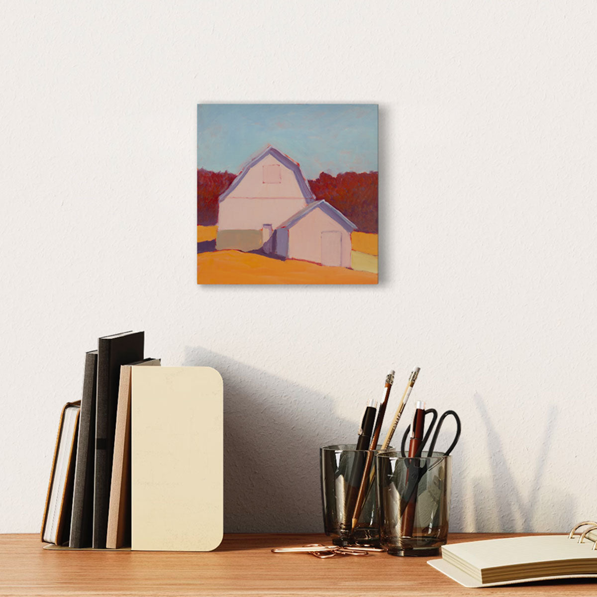 A pink and red contemporary barn landscape painting hangs on a white wall above a desk with book, glasses holding pens, and an open notebook. 