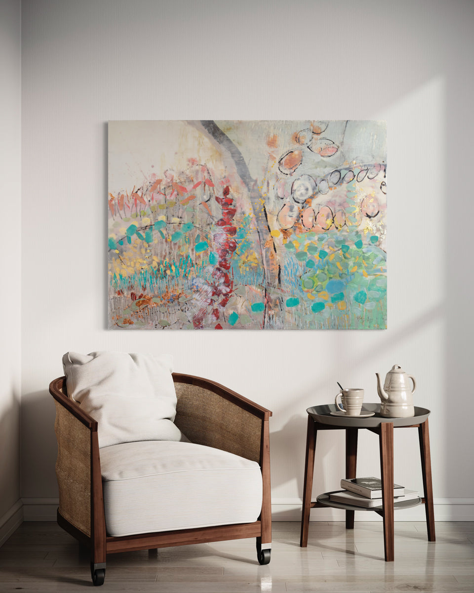 An abstract multicolored painting hangs on a white wall above a wood armchair with white cushions and a modern end table with ceramic teapot and teacup. 