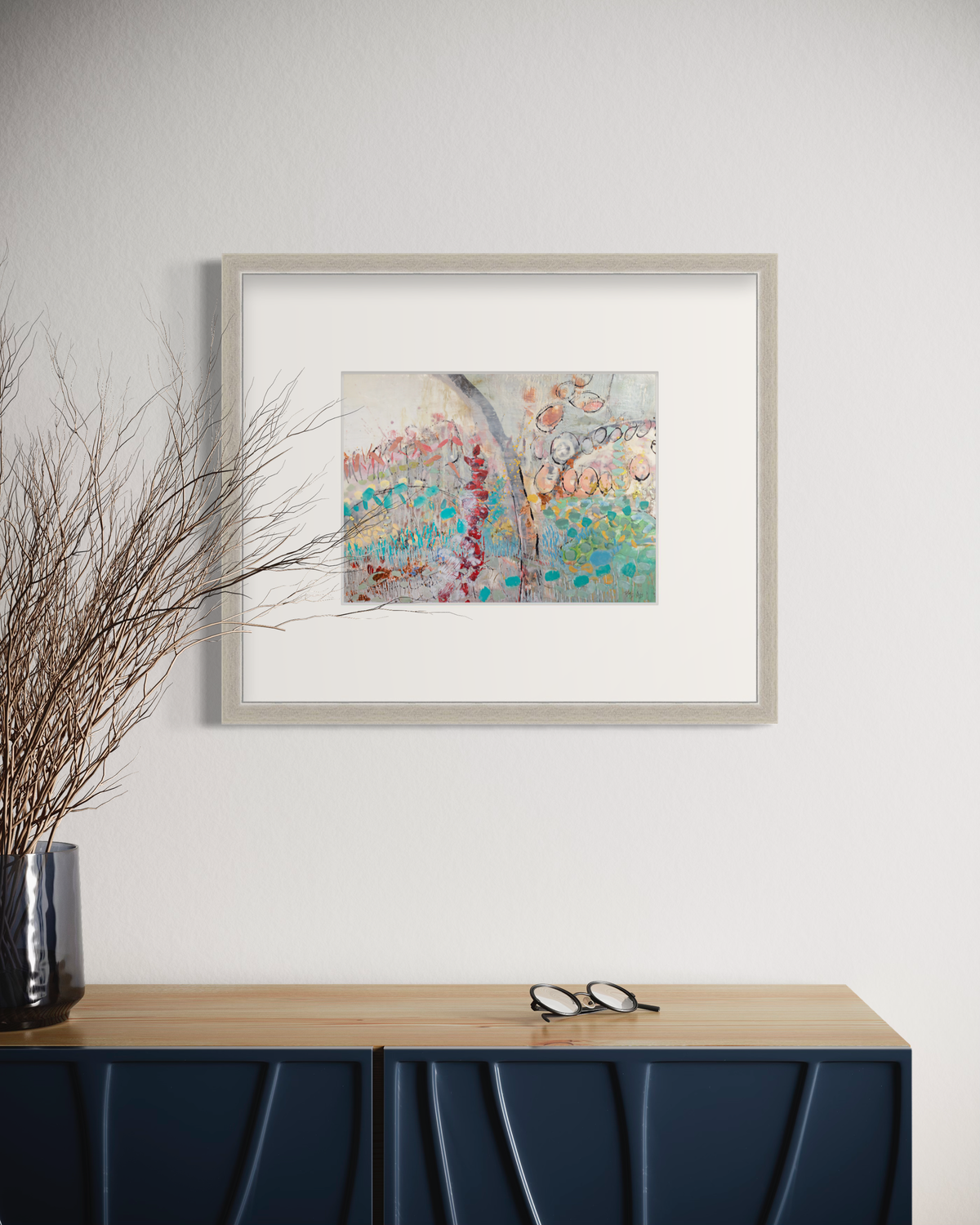 A multicolored abstract floral print in a silver frame with a mat hangs on a white wall above a navy blue and wood cabinet with a pair of eyeglasses and a base with wood stems.