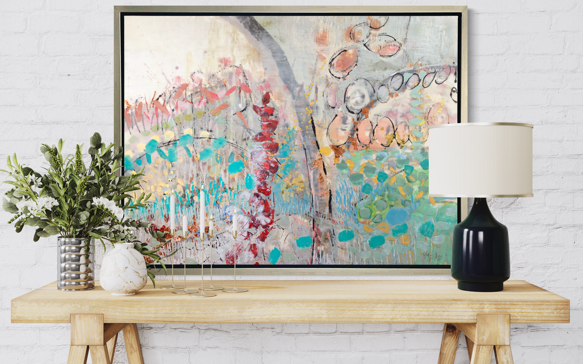 A multicolored abstract floral print in a silver floater frame hangs on a white brick wall above a raw wood table with a table lamp, five tapered candles, a white marble vase and a chrome can vase with greenery.
